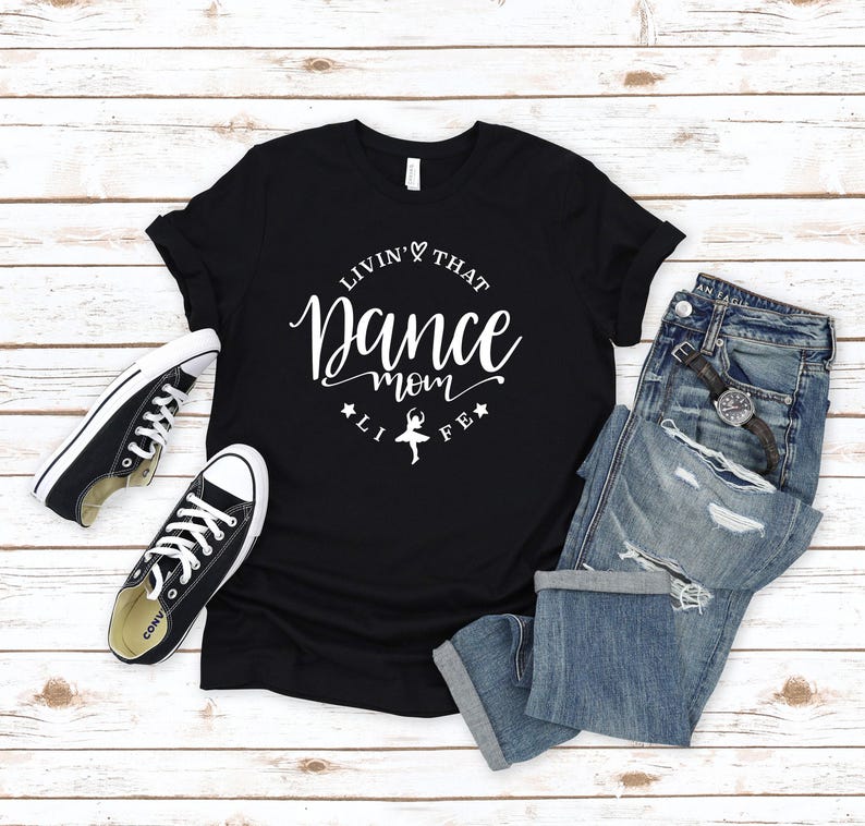 A black t-shirt with white text, a pair of black and white sneakers, and a pair of blue jeans are laid out on a wooden surface.