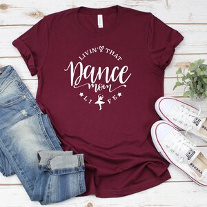A burgundy t-shirt with the text "LIVIN' THAT DANCE MOM" printed on it, along with a pair of blue jeans and white sneakers, all laid out on a wooden surface.