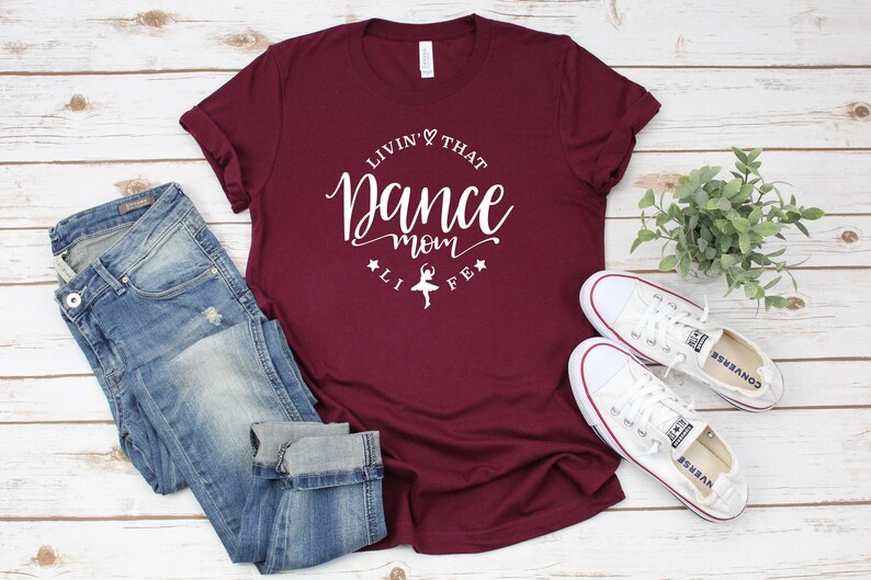 A burgundy t-shirt with the text "LIVIN' THAT DANCE MOM" printed on it, along with a pair of blue jeans and white sneakers, all laid out on a wooden surface.