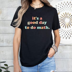 May include: Black t-shirt with the text "it's a good day to do math" in colourful letters. The shirt is worn by a person wearing blue jeans and bracelets.