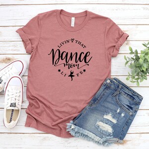A pink t-shirt with a black text design, a pair of denim shorts, and a pair of white sneakers are laid out on a wooden surface.