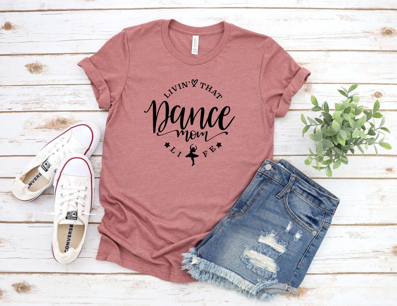 A pink t-shirt with a black text design, a pair of denim shorts, and a pair of white sneakers are laid out on a wooden surface.