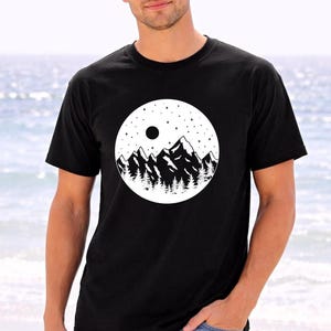A man wearing a black t-shirt with a graphic design of mountains and a moon on it, standing in front of a body of water.