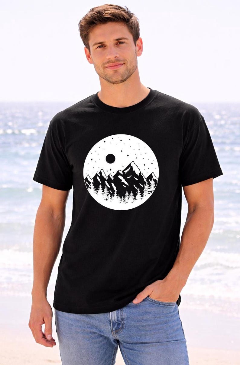 A man wearing a black t-shirt with a graphic design of mountains and a moon on it, standing in front of a body of water.