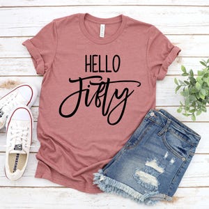 A pink t-shirt with the text "Hello Fifty" printed on it, a pair of denim shorts, and a pair of white sneakers are laid out on a wooden surface.
