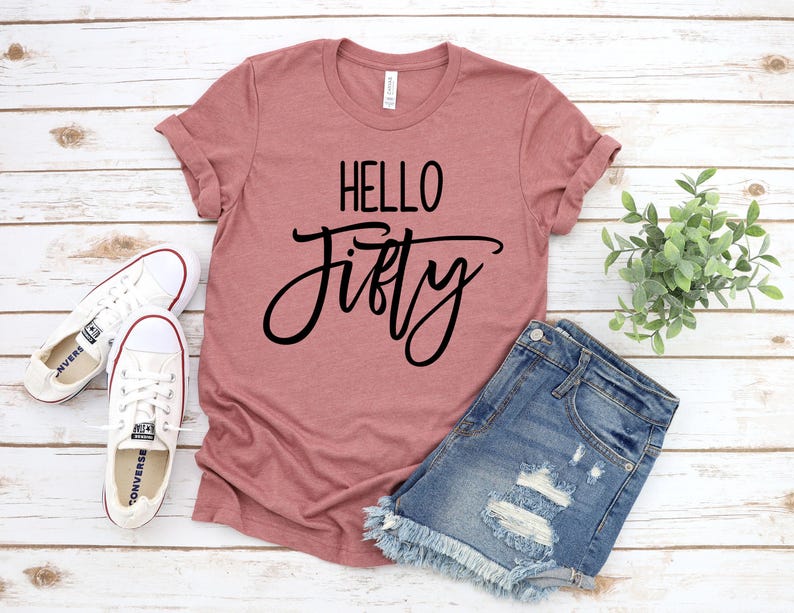 A pink t-shirt with the text "Hello Fifty" printed on it, a pair of denim shorts, and a pair of white sneakers are laid out on a wooden surface.