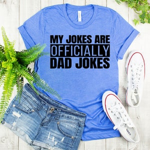 K&ouml;nnte beinhalten: Ein blaues T-Shirt mit dem Text "My Jokes Are Officially Dad Jokes" in Schwarz.