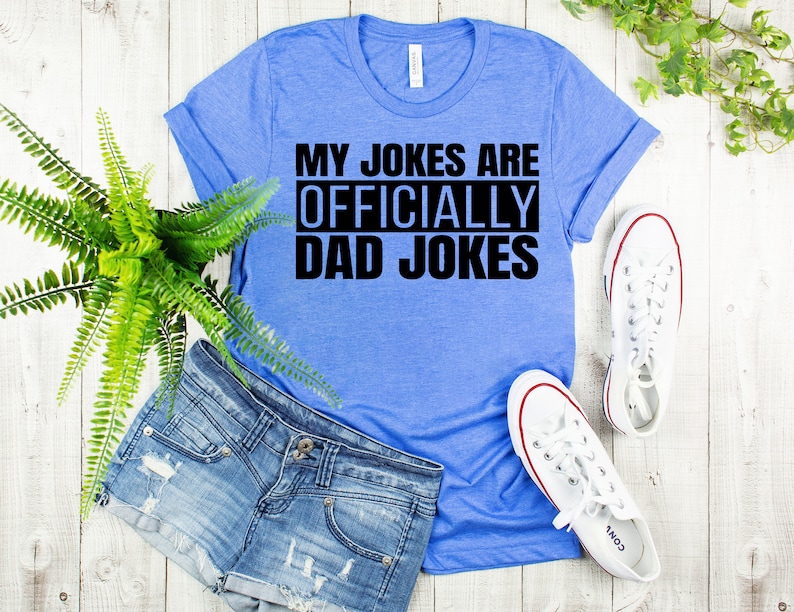 K&ouml;nnte beinhalten: Ein blaues T-Shirt mit dem Text "My Jokes Are Officially Dad Jokes" in Schwarz.