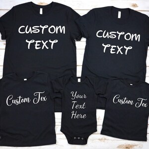 Peut inclure: Plusieurs t-shirts noirs et un body de bébé. Ils affichent du texte blanc : « Custom Text » et « Your Text Here ». Les t-shirts sont à manches courtes et à col rond. Le body a des boutons-pression en bas.