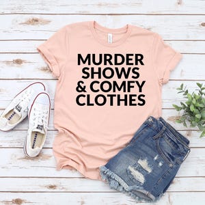A pink t-shirt with the text "MURDER SHOWS & COMfy CLOTHES" printed on it, along with a pair of denim shorts and white sneakers, all laid out on a wooden surface.