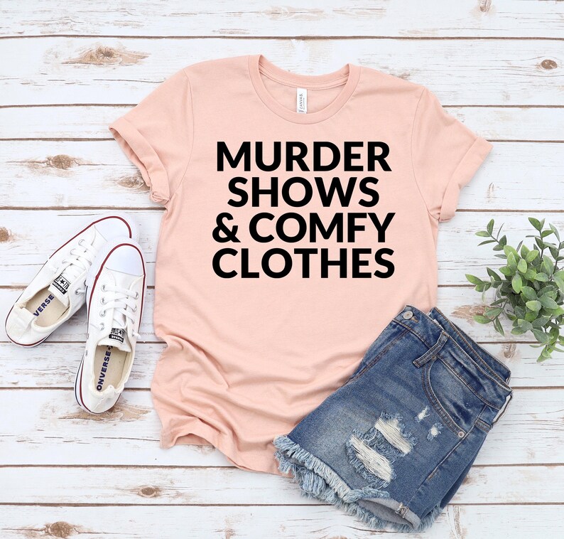 A pink t-shirt with the text "MURDER SHOWS & COMfy CLOTHES" printed on it, along with a pair of denim shorts and white sneakers, all laid out on a wooden surface.