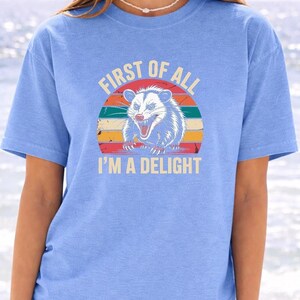 A woman wearing a blue t-shirt with a graphic design of a smiling opossum and the text "First of all I'm a delight".