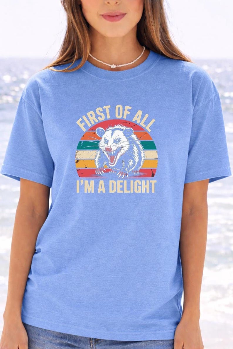 A woman wearing a blue t-shirt with a graphic design of a smiling opossum and the text "First of all I'm a delight".