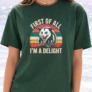 A woman wearing a green t-shirt with a graphic design of a smiling opossum and the text "First of all I'm a delight".