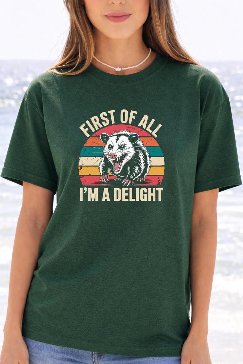 A woman wearing a green t-shirt with a graphic design of a smiling opossum and the text "First of all I'm a delight".