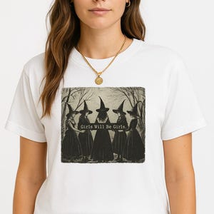 May include: White t-shirt featuring a vintage-style illustration of five figures in witch hats and dresses. The text "Girls Will Be Girls" is printed below the image. The shirt is made of a soft material and has short sleeves.