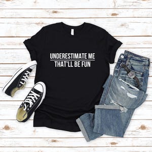 May include: Black t-shirt with white text that reads "UNDERESTIMATE ME THAT'LL BE FUN". The shirt is laid flat on a white wood surface with a pair of black sneakers and blue jeans.