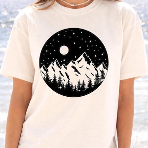 A woman wearing a white t-shirt with a graphic design of a mountain landscape under a night sky with a full moon and stars.