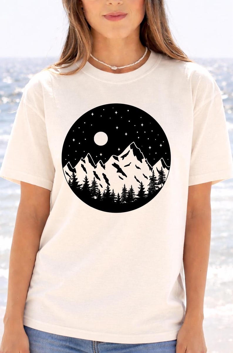 A woman wearing a white t-shirt with a graphic design of a mountain landscape under a night sky with a full moon and stars.
