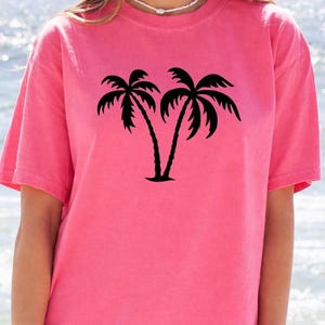 A woman wearing a pink t-shirt with a graphic of two palm trees printed on it.
