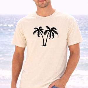 A young man wearing a white t-shirt with a graphic of two palm trees stands on a beach, with the ocean visible in the background.