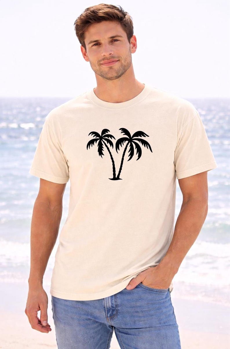 A young man wearing a white t-shirt with a graphic of two palm trees stands on a beach, with the ocean visible in the background.