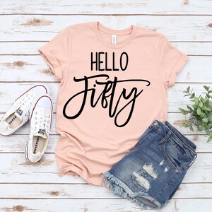 A pink t-shirt with the text "HELLO FIFTY" printed on it, along with a pair of denim shorts and white sneakers, all laid out on a wooden surface.