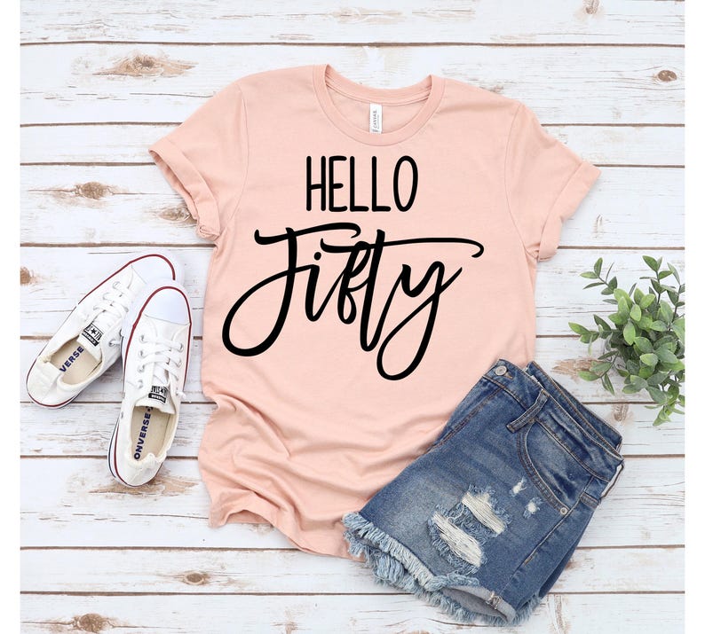 A pink t-shirt with the text "HELLO FIFTY" printed on it, along with a pair of denim shorts and white sneakers, all laid out on a wooden surface.