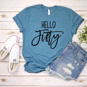 A blue t-shirt with the text "Hello Fifty" printed on it, along with a pair of denim shorts and white sneakers, all laid out on a wooden surface.