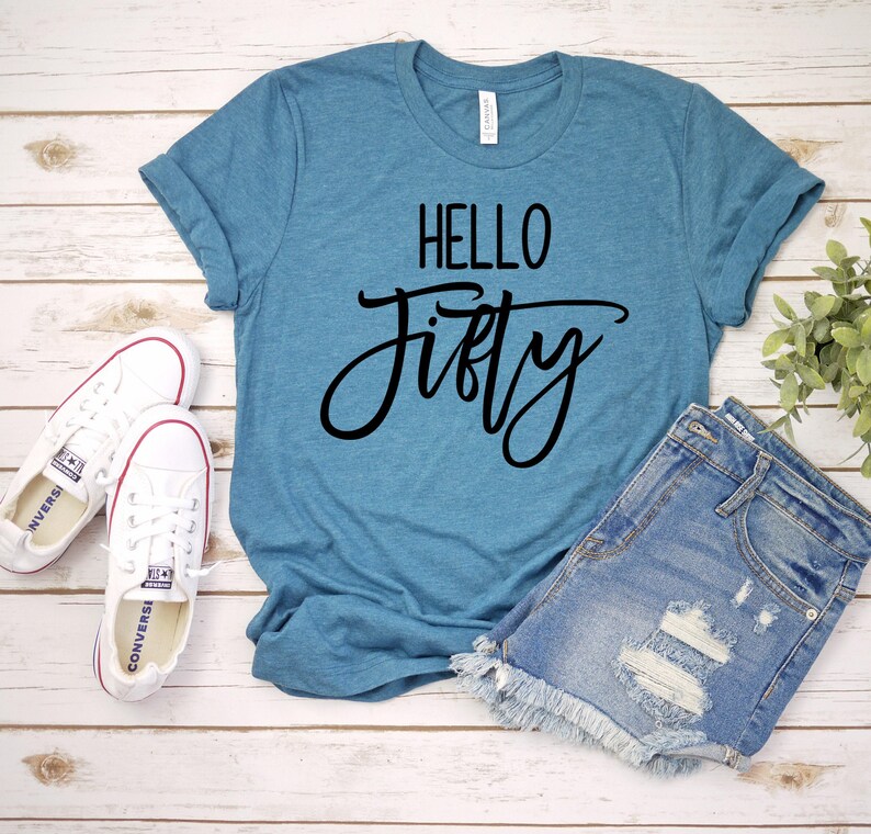 A blue t-shirt with the text "Hello Fifty" printed on it, along with a pair of denim shorts and white sneakers, all laid out on a wooden surface.