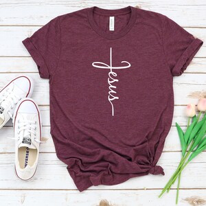 K&ouml;nnte beinhalten: Ein maroonfarbenes T-Shirt mit einer wei&szlig;en Grafik eines Kreuzes, auf dem das Wort "Jesus" steht. Das Shirt liegt auf einer wei&szlig;en Holzoberfl&auml;che mit einem Paar wei&szlig;en Turnschuhen und einem Strau&szlig; rosa Tulpen.