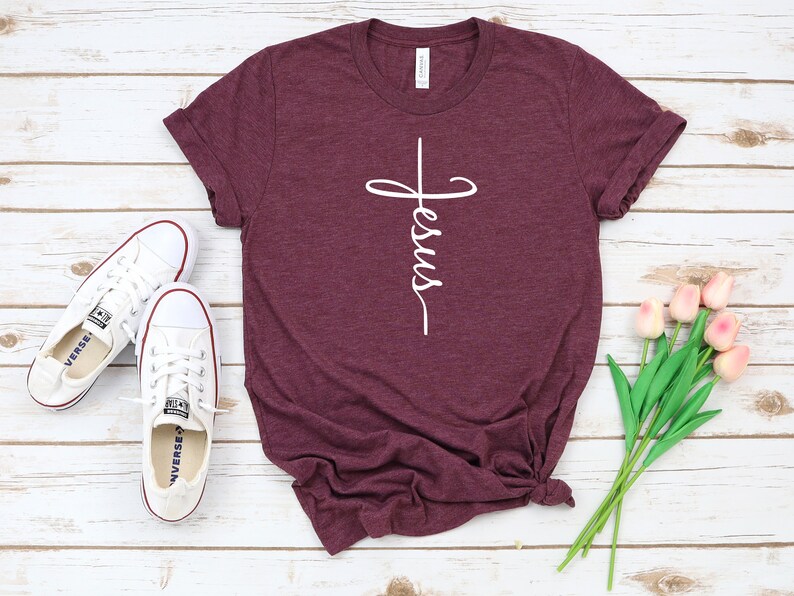 K&ouml;nnte beinhalten: Ein maroonfarbenes T-Shirt mit einer wei&szlig;en Grafik eines Kreuzes, auf dem das Wort "Jesus" steht. Das Shirt liegt auf einer wei&szlig;en Holzoberfl&auml;che mit einem Paar wei&szlig;en Turnschuhen und einem Strau&szlig; rosa Tulpen.