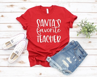 Könnte beinhalten: Ein rotes T-Shirt mit dem Text "Santa's Favorite Teacher" in weißer Schrift. Das Shirt wird mit Jeansshorts und weißen Turnschuhen kombiniert. Eine kleine grüne Pflanze befindet sich ebenfalls im Bild, alles auf einer hellen Holzoberfläche.