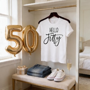 a 50th birthday celebration, with a white t-shirt hanging on a hanger that says "Hello Fifty", a pair of jeans, a pair of sneakers, and a gift box. There are also gold foil balloons shaped like the number 50, and a glass of champagne on a table.