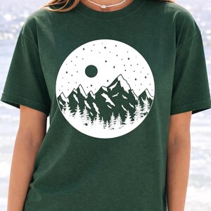A woman wearing a green t-shirt with a white graphic design depicting a mountain landscape with a full moon and stars.