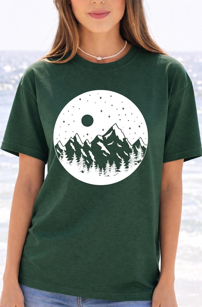 A woman wearing a green t-shirt with a white graphic design depicting a mountain landscape with a full moon and stars.