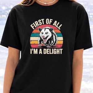 A woman wearing a black t-shirt with a graphic design of a smiling opossum and the text "First of all I'm a delight".