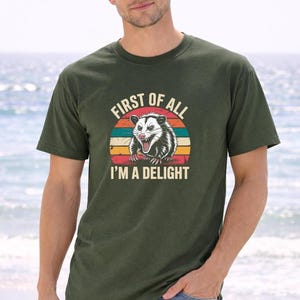 A man wearing a green t-shirt with a graphic of a smiling opossum and the text "First of all I'm a delight" standing on a beach.