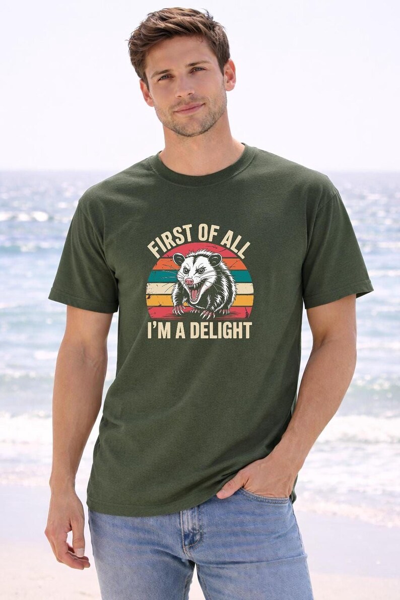 A man wearing a green t-shirt with a graphic of a smiling opossum and the text "First of all I'm a delight" standing on a beach.