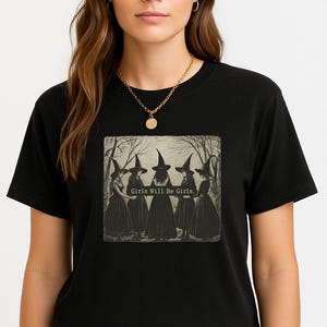 May include: Black t-shirt featuring a vintage-style graphic of five witches in long dresses and pointed hats. The text "Girls Will Be Girls" is printed below the image. The model is wearing a gold necklace.