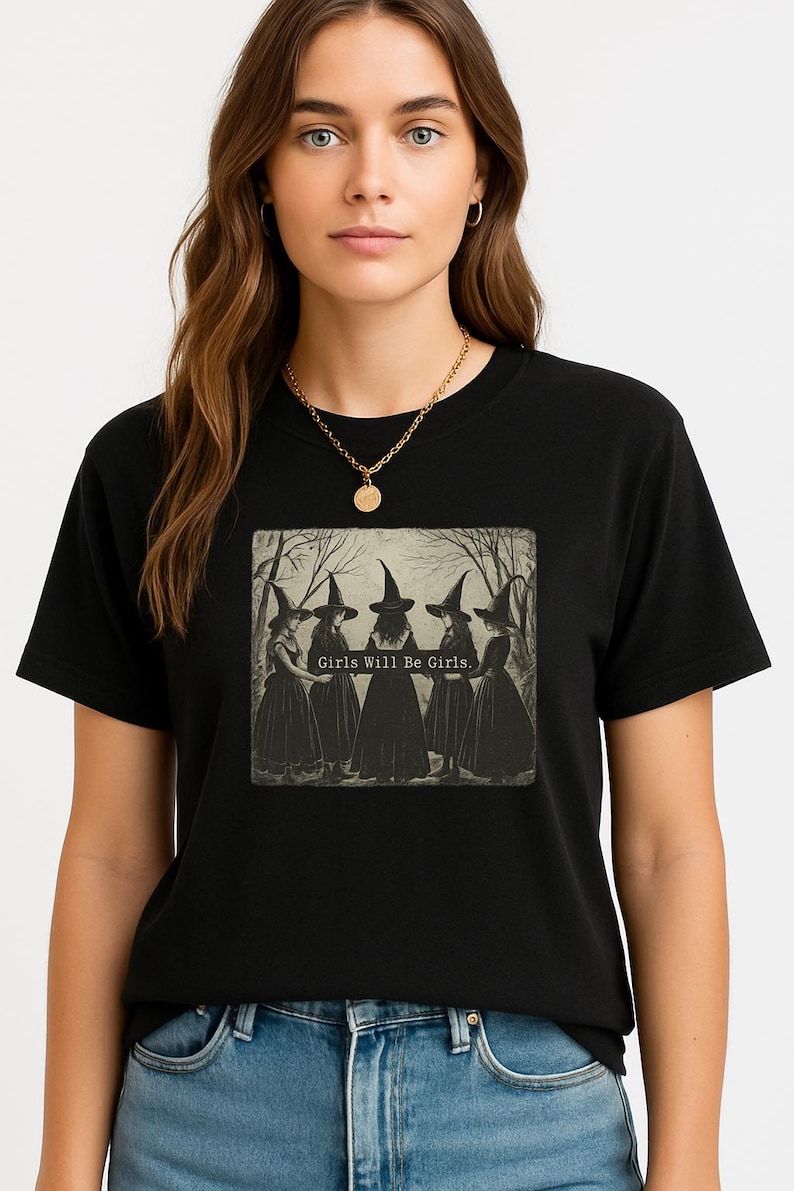 May include: Black t-shirt featuring a vintage-style graphic of five witches in long dresses and pointed hats. The text "Girls Will Be Girls" is printed below the image. The model is wearing a gold necklace.