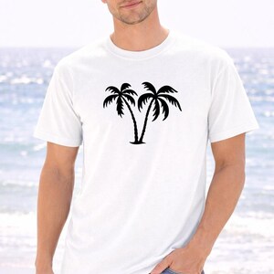 A man wearing a white t-shirt with a graphic of two palm trees stands on a beach, with the ocean visible in the background.