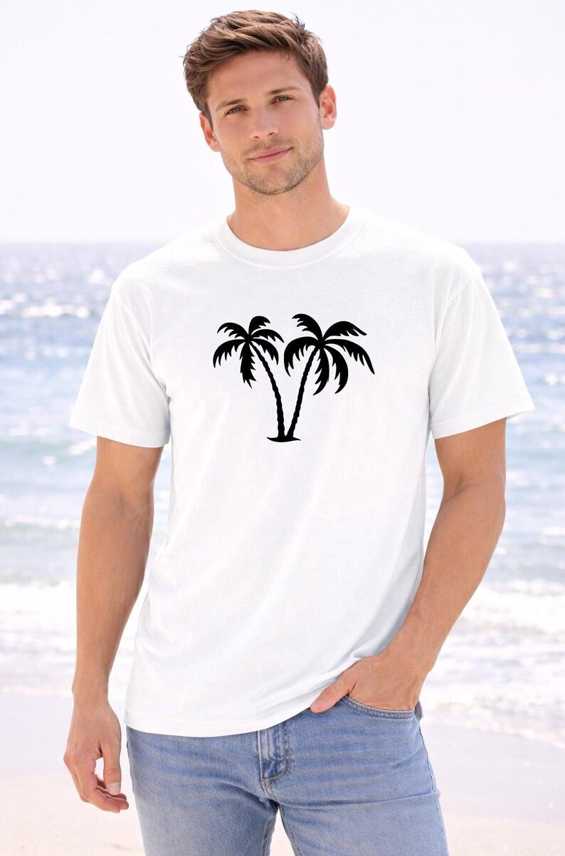 A man wearing a white t-shirt with a graphic of two palm trees stands on a beach, with the ocean visible in the background.