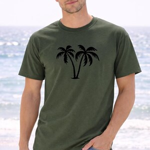 A young man wearing a green t-shirt with a palm tree graphic stands on a beach, with the ocean visible in the background.