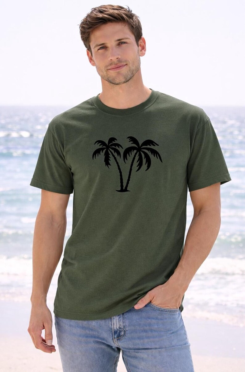 A young man wearing a green t-shirt with a palm tree graphic stands on a beach, with the ocean visible in the background.