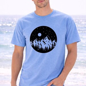 A man wearing a blue t-shirt with a mountain landscape design stands in front of a body of water.