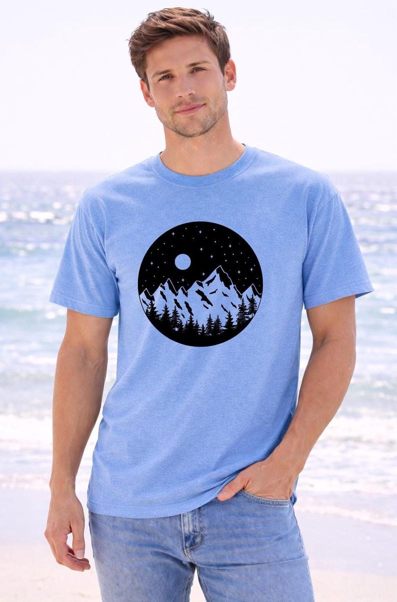A man wearing a blue t-shirt with a mountain landscape design stands in front of a body of water.