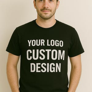 May include: Black crew neck t-shirt with the text "YOUR LOGO CUSTOM DESIGN" in white. The shirt is a solid black colour and has short sleeves. The design is centered on the front of the shirt.