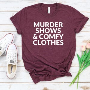 a burgundy t-shirt with the text "MURDER SHOWS & COMfy CLOTHES" printed on it, along with a pair of white sneakers and a bouquet of pink flowers.