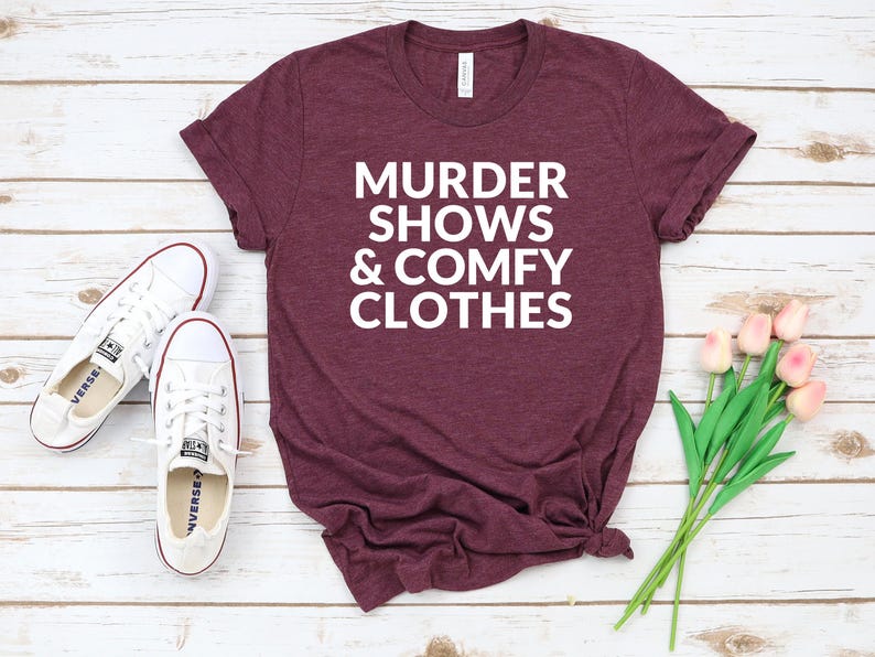 a burgundy t-shirt with the text "MURDER SHOWS & COMfy CLOTHES" printed on it, along with a pair of white sneakers and a bouquet of pink flowers.